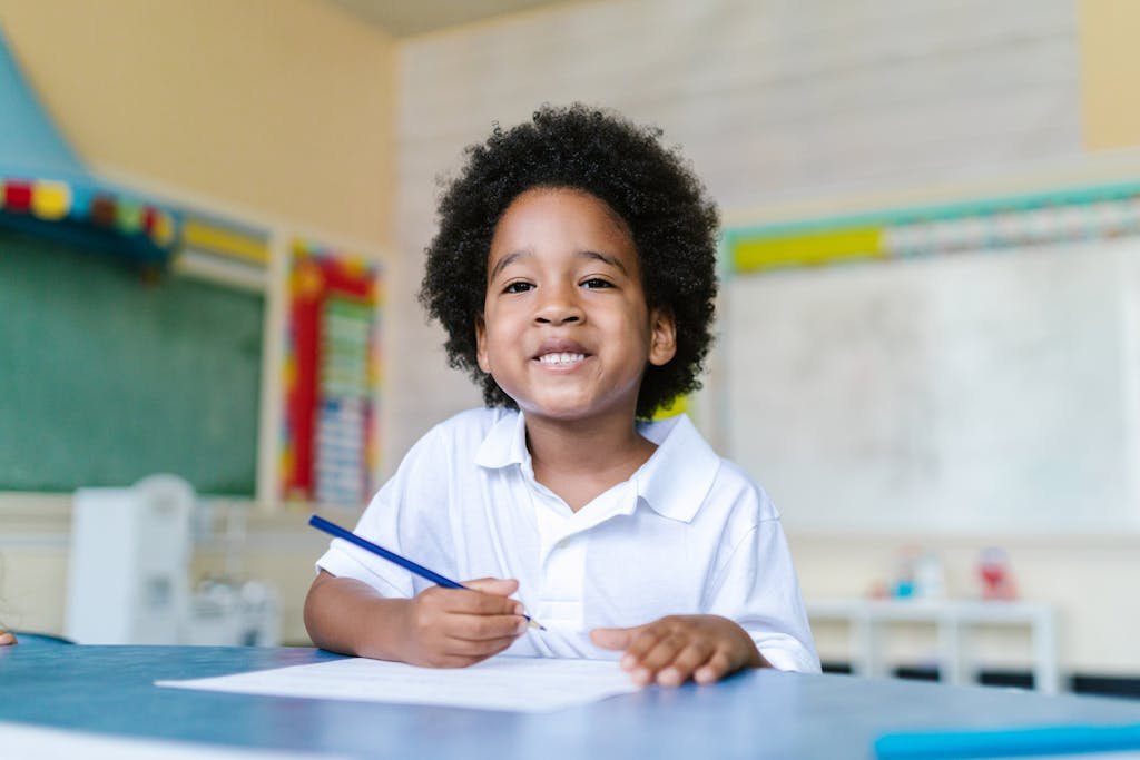 Happy child with afro hair sitting in a classroom, holding a pencil and smiling.